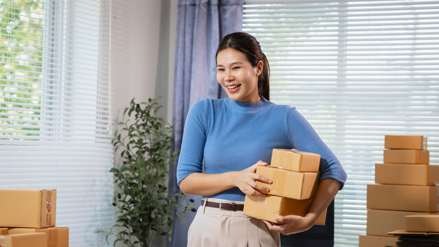 Smiling young woman holding small cardboard boxes in a bright home office, surrounded by packages, suggesting online selling, packing, delivery, and small business work.