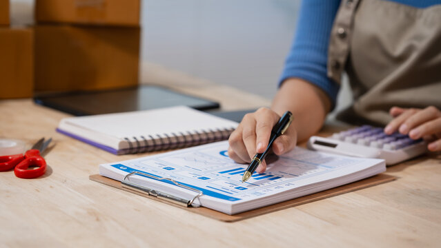 Person writing on a clipboard form while using a calculator at a desk, with notebook and boxes nearby, showing small business bookkeeping, budgeting, and home office planning.