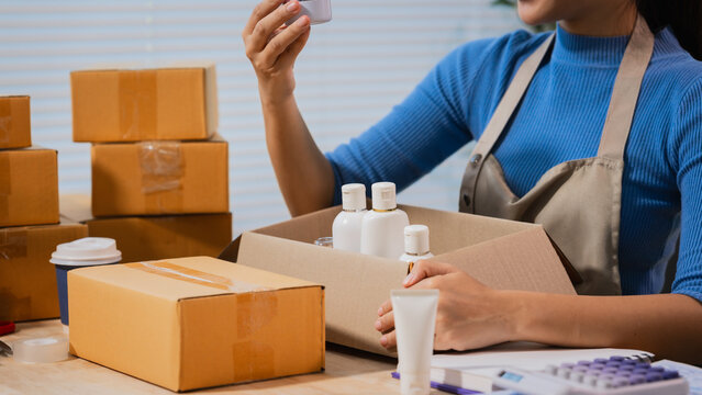 Woman packing skincare bottles into shipping boxes at a home workspace, checking a product while preparing online orders for small business delivery.