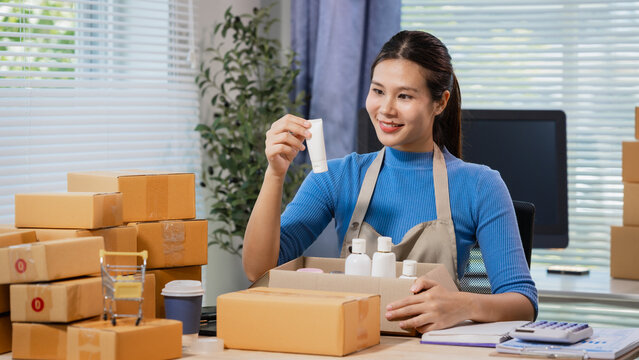 Young woman packing beauty products in cardboard boxes at a home office, checking a label for online orders. Clean workspace suggests small business, shipping, retail, and entrepreneurship.