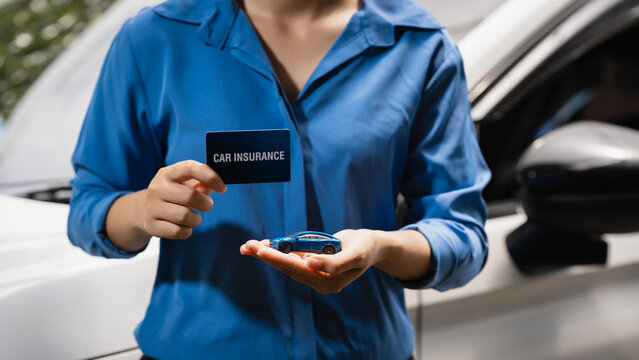 Woman in blue shirt holding a car insurance card and small toy car beside a white vehicle, showing auto coverage, safety, and protection for drivers.