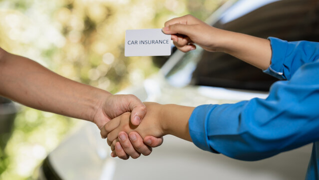 Close-up of two people shaking hands beside a car while one holds an insurance card, showing agreement, support, and vehicle coverage after an accident or claim.