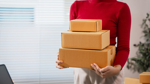 Woman in a red top holding a stack of cardboard shipping boxes indoors, showing delivery, online shopping, packing, moving, or small business order fulfillment.