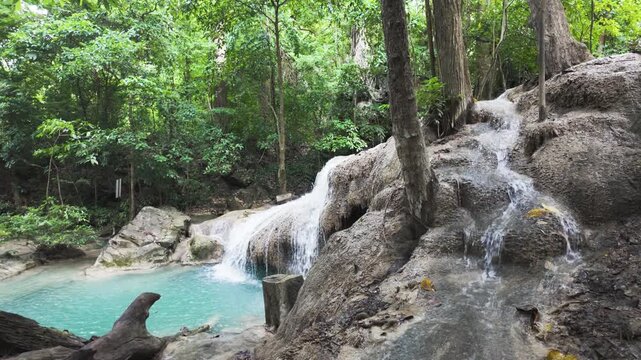 Clear stream water slips over textured rock tiers into an aqua pool in a lush Kanchanaburi woodland, with a gentle rack focus shift.