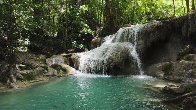 A school of fish glides through a blue-green lagoon at the base of a tiered waterfall framed by lush Thai vegetation.