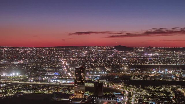 A panoramic aerial view of a sprawling city at dusk, with an orange to purple gradient in the sky, city lights coming on, mountains in the background, and a cinematic time-lapse.