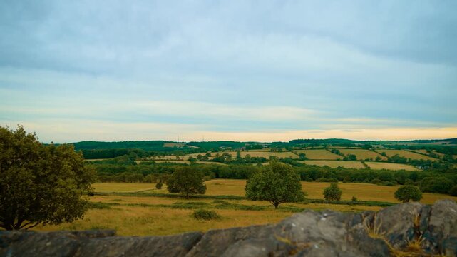 Rolling countryside valley beneath layered cloudscape. Green pasture fields with scattered trees distant hills and agricultural farmland panorama. Rural summer landscape with open meadows cultivated