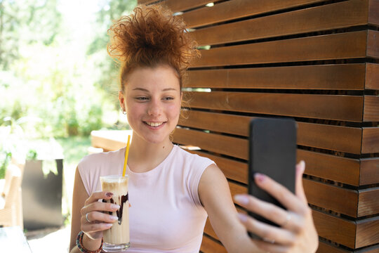 A young woman taking a selfie with a mobile phone, drinking coffee in a cafe, and smiling