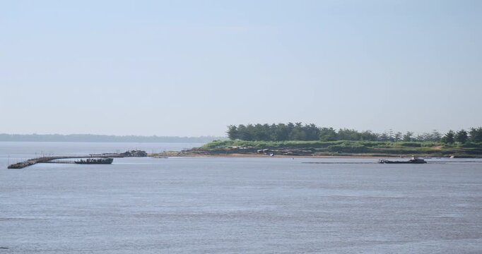 River flows past a island dotted with trees, featuring a ferry packed with people docking at a bamboo bridge under construction, while another boat assists with the bridge building in the distance