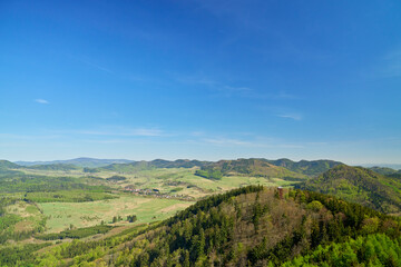 Fototapeta premium Panoramic view of green hills, forested slopes, open valley and small lookout tower under blue sky. Concept of nature travel, hiking and rural landscape.