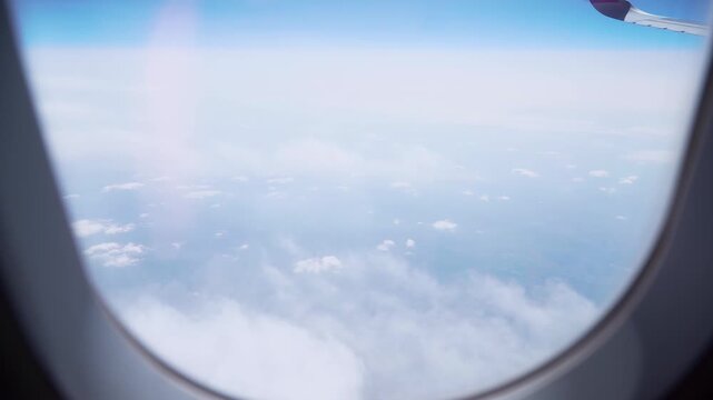 Airplane flying above clouds with view through passenger window and wing visible in distance. Window frame and glass edges surrounding sky and cloud layer below. Air travel journey, aviation transport