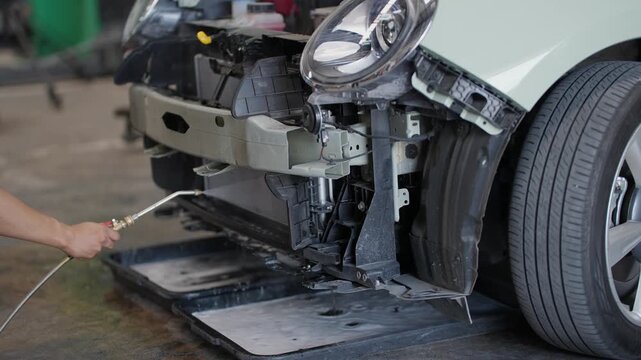 EV Technician cleans car's front bumper with compressed air. Vehicle sits on lift, ready for repair. Workshop environment suggests routine maintenance. No people visible in shot