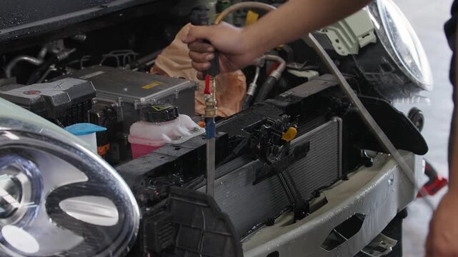 EV Mechanic cleans engine bay with spray. Steam rises from car's undercarriage. Hand holds tool near coolant reservoir. Engine compartment is open and cluttered. Scene shows routine maintenance work