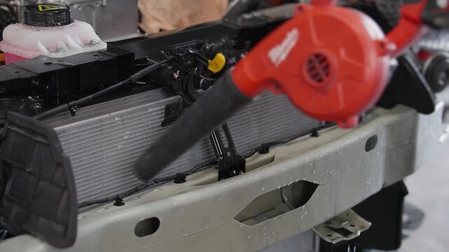 Red blower clears debris from EV car radiator. Mechanic works carefully on vehicle cooling system. Blower directs air to clean heat exchanger. Perfect for auto repair or maintenance projects