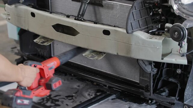 Worker clears debris from EV car radiator with handheld blower. Hands grip tool firmly during maintenance task. Vehicle front is exposed, radiator is detached