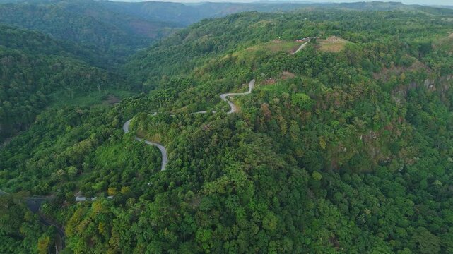 Scenic drone footage capturing a curvy, serpentine asphalt road cutting through dense tropical forest and rolling hills at Geopark Ciletuh, Sukabumi, Indonesia