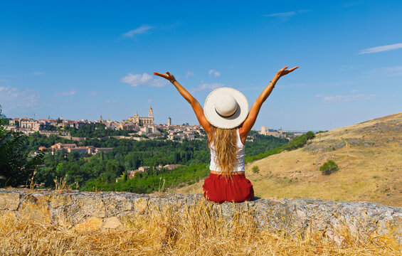 Rear view of happy young woman tourist with open arms celebrating on stone wall looking at panoramic skyline of Segovia Spain