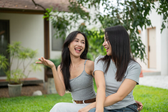 Two cheerful Asian female friends chatting and laughing together on the green grass lawn in front of a modern house.