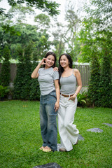 Fototapeta premium Asian female friends standing on grass side by side and smiling in a beautiful greenery garden cafe.