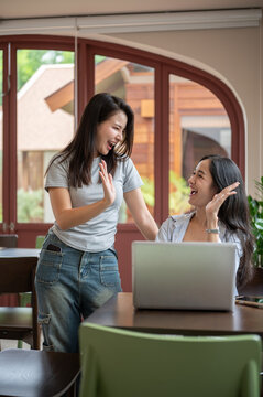 Asian women coworkers smiling while doing a high five together over laptop at table in a bright cafe