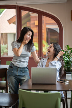 Asian women friends looking surprised while celebrating teamwork and success beside a laptop in cafe