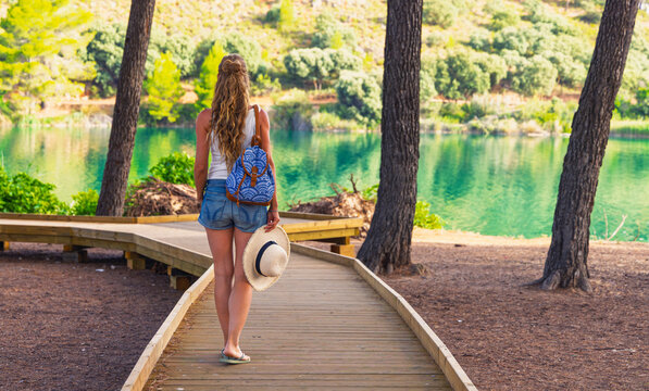 Rear view of young woman with backpack and straw hat walking on wooden pier by beautiful turquoise lake in pine forest