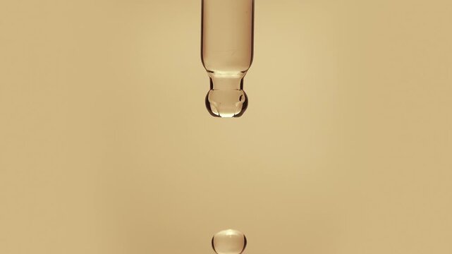 Macro shot of a golden vitamin C ascorbic acid or argan oil droplet falling from a glass pipette dropper isolated on a solid beige background