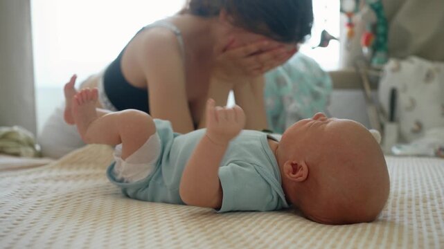Depressed young mother experiencing postpartum depression, covering her face with her hands in desperation while her newborn baby is crying continuously on the bed in the bedroom