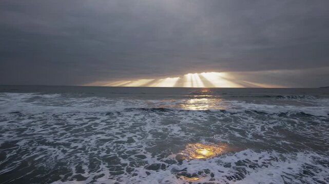 Ocean waves crash against the shore under a dramatic sky with rays of sunlight breaking through the clouds, creating a serene coastal atmosphere