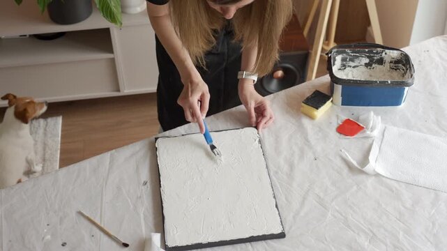Female artist shapes relief texture on canvas with white plaster paste and palette knife at a covered work table. Concept of DIY wall decor and creative hobby.