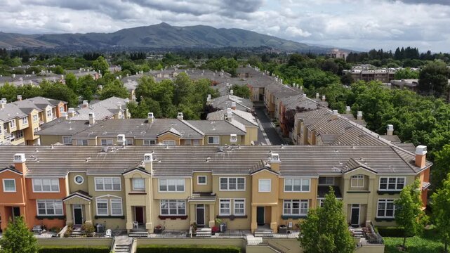 Colorful multi-family townhomes line quiet Fremont, California street, long rows of parked cars filling curbside spaces beneath tree-covered rooftops near hazy mountains. Aerial drone approaching shot