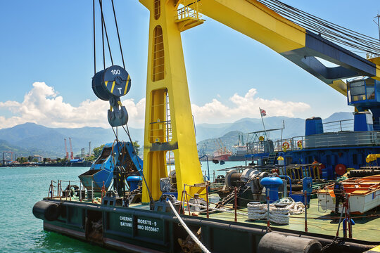 Batumi, Georgia - July 1, 2026: Heavy floating crane barge deck equipment, yellow steel boom, lifting hook, winch closeup docked in harbor against mountains.