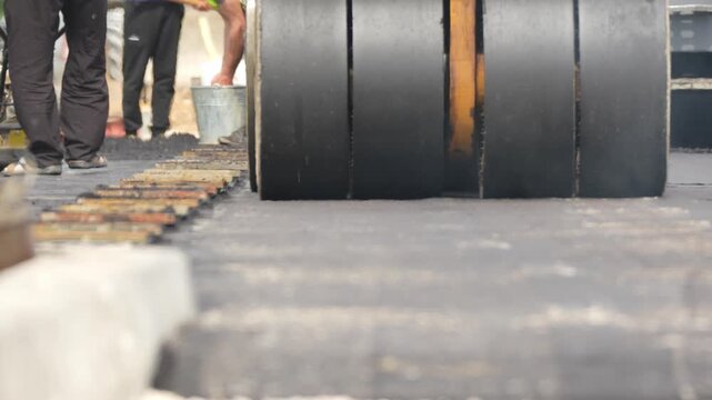 Black paving roller stands on concrete. Workers move paving stones with shovels. They concentrate on road construction. Ideal for construction footage. Show urban development efforts