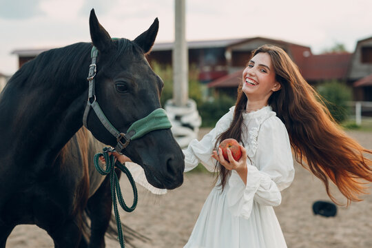 Young smiling woman feeds her horse an apple at outdoors ranch stable