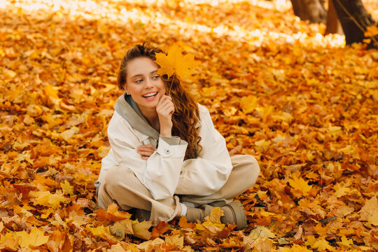 Smiling young woman sits in the autumn foliage ground  on the forest covered yellow leaves.