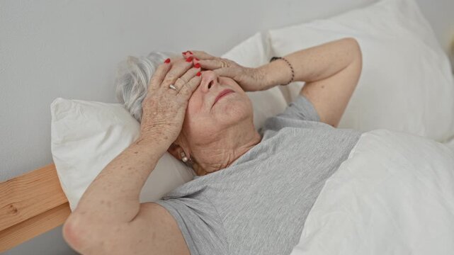 Woman holds forehead with hands on bed inside a building, covering eyes and signaling headache; fatigue vulnerability.