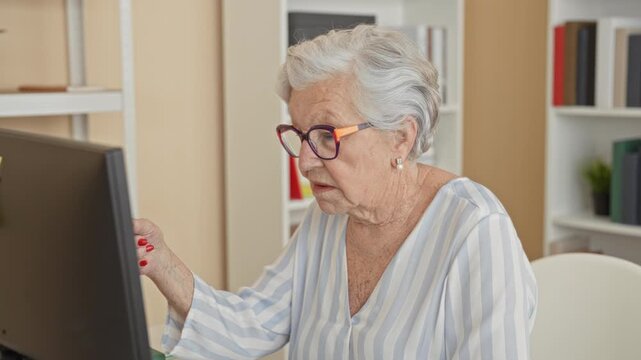 Senior woman points index finger at computer monitor, hand visible, wearing glasses in a building office; concentration independence.