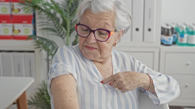 Woman points finger to bandage on upper arm in clinic building while wearing striped blouse and purple glasses; relief trust.