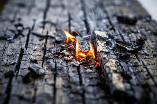 Small flames flickering on charred black wood surface with wisps of smoke rising from dark embers.