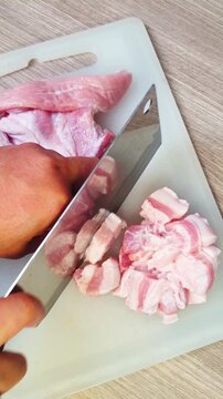 A person using a kitchen cleaver knife to chop raw pork belly into pieces on a white cutting board