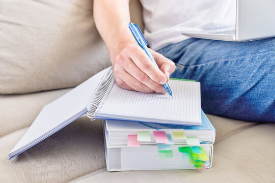 Close-up of student writing notes in notebook while sitting on sofa at home, study and education concept for homework, exam preparation, remote learning, planning, productivity and academic routine
