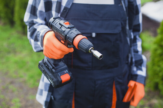 person holding power drill outdoors wearing orange gloves and black jacket for home improvement project in garden setting close-up practical and serene mood natural light clean background.