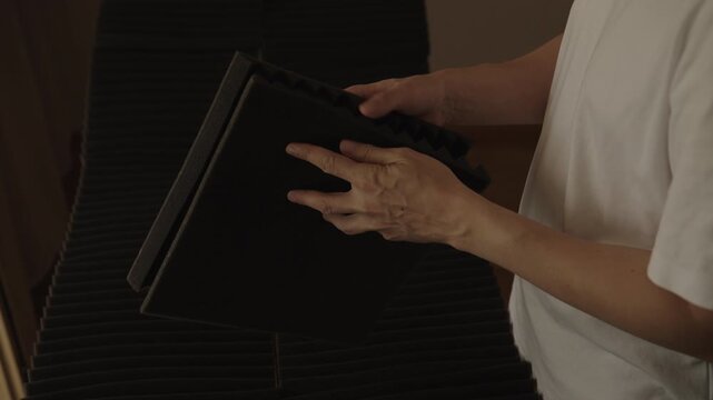 Cropped shot of man in white shirt handles and stacks black acoustic foam panels. Preparing for soundproofing room, home studio setup, acoustic treatment project. DIY sound absorption.