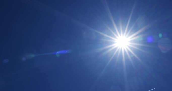 Bright starburst sun moving across a clear blue sky with beautiful shifting lens flares, slow camera pan left