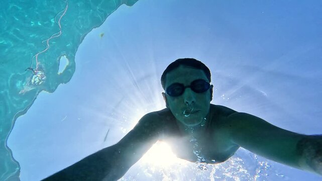 Slow-motion of a man swimming underwater in the Mediterranean Sea. The man is of Caucasian origin, is forty-two years old, has short hair and has swimming glasses on his face