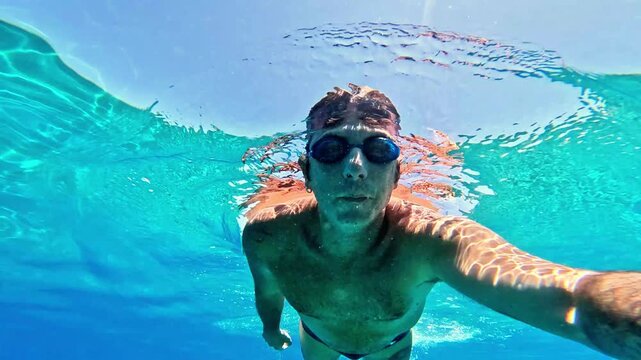 Slow-motion of a man swimming underwater in the Mediterranean Sea. The man is of Caucasian origin, is forty-two years old, has short hair and has swimming glasses on his face