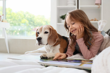Obraz premium Cute little girl with Beagle dog reading book on bed in bedroom