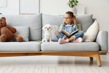 Obraz premium Cute little happy girl with Maltese dog reading book on sofa at home