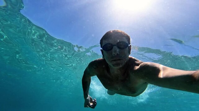 Slow-motion of a man swimming underwater in the Mediterranean Sea. The man is of Caucasian origin, is forty-two years old, has short hair and has swimming glasses on his face