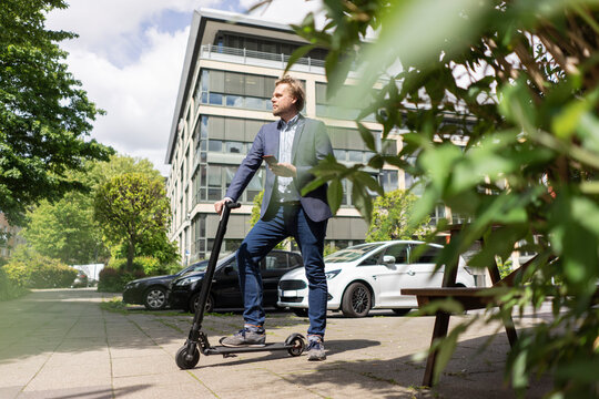Businessman standing with E-Scooter and smartphone on pavement in the city